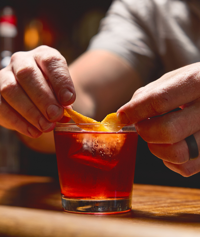 A person garnishing a dark red cocktail with a lemon twist, placed on a wooden surface.