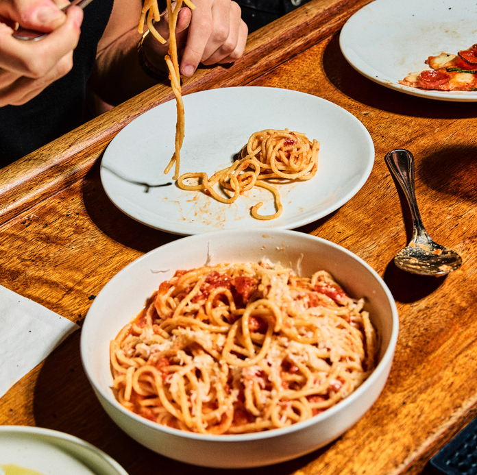 Person eating spaghetti with tomato sauce at a wooden table with plates and utensils.