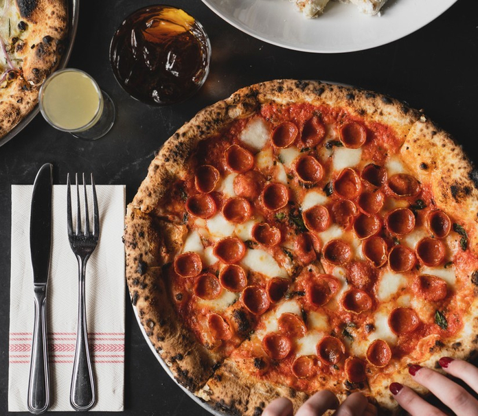 A close-up of a pepperoni pizza on a dark table, with a glass of beer and another pizza in the background.