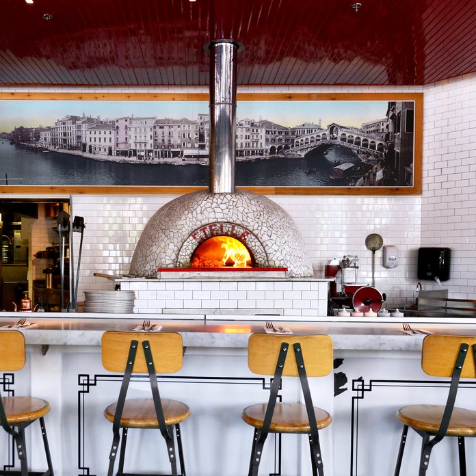 Modern pizzeria interior with red ceiling, white subway tile backsplash, black iron bar stools, and a large, arched pizza oven emitting light.
