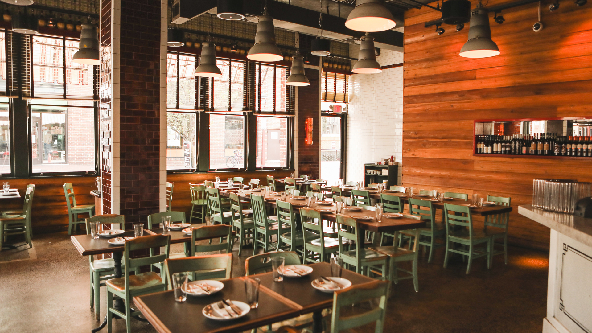 Modern restaurant with wooden tables, green chairs, and warm lighting. Large windows let in natural light. Wall display of beverages in background.