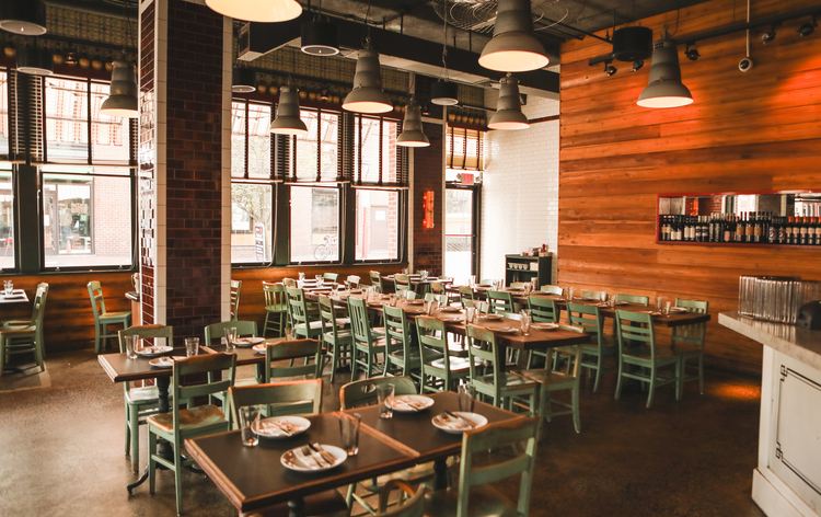 Modern restaurant interior with wooden walls, industrial pendant lights, and tables with green chairs arranged for dining.