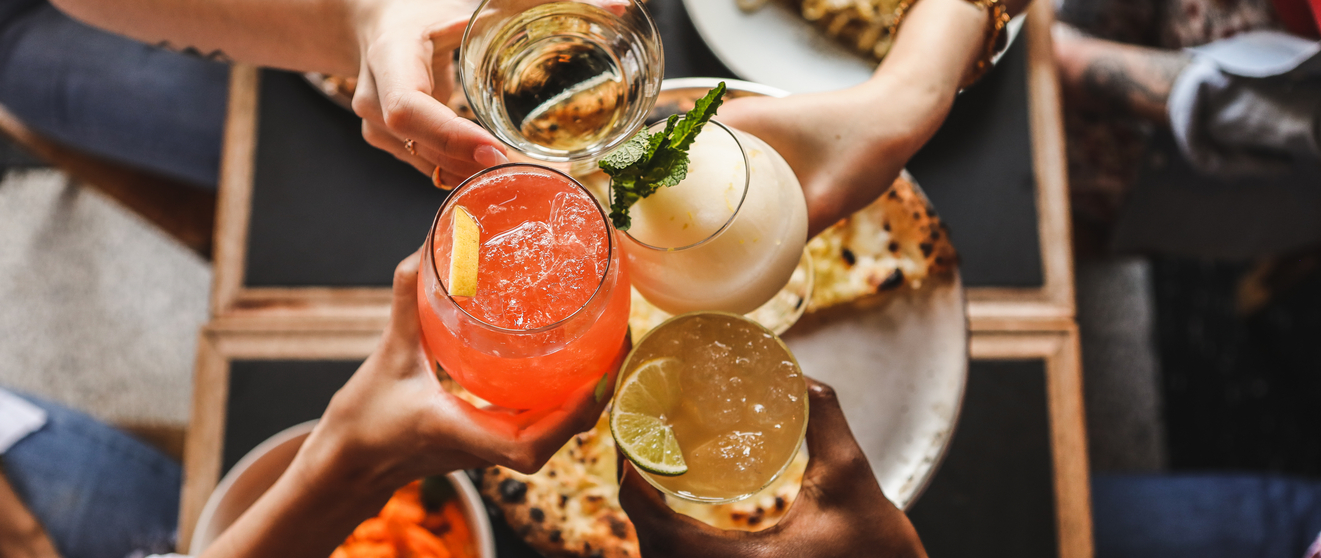 Group toasting with drinks and plates of pizza and pasta on a wooden table.