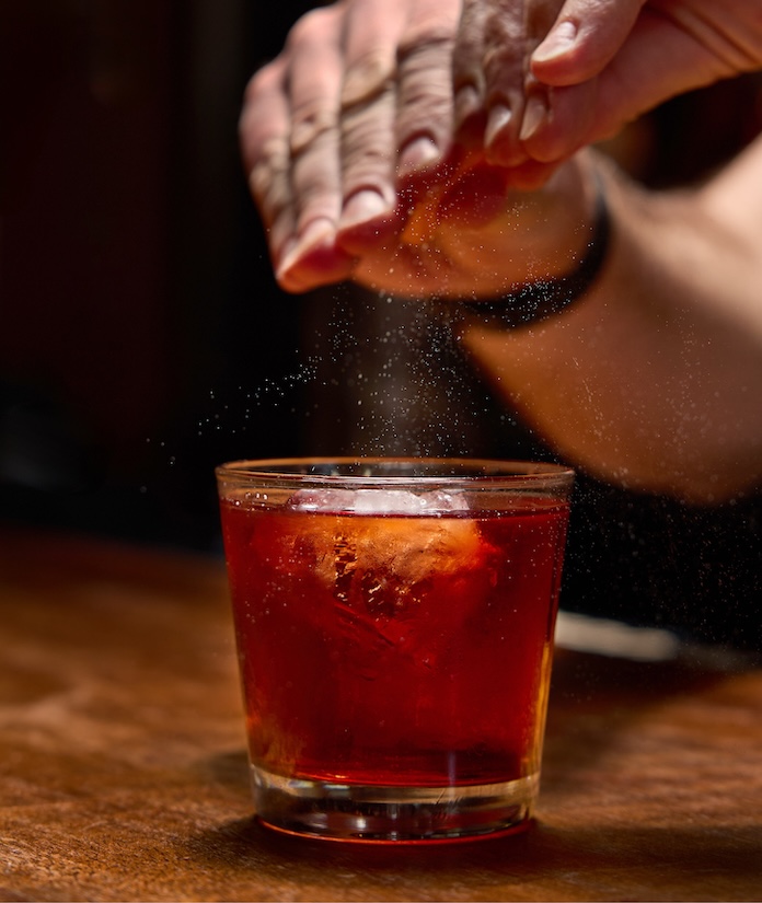 A person garnishing a dark red cocktail with a lemon twist, placed on a wooden surface.
