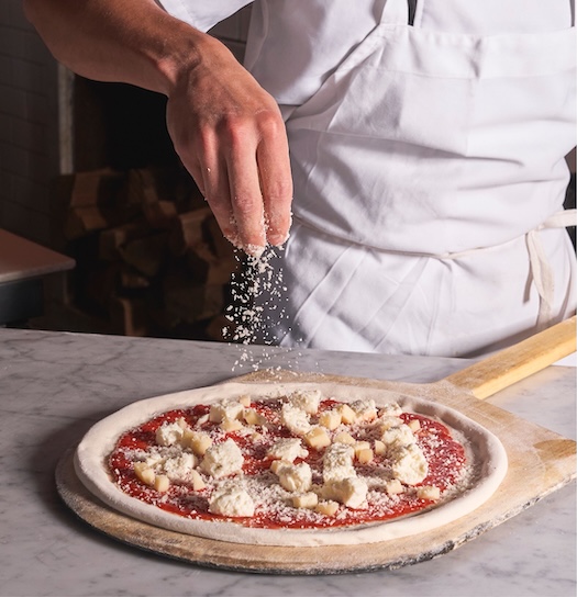 Chef preparing a Neapolitan pizza, sprinkling grated cheese over fresh mozzarella and tomato sauce on a wooden peel