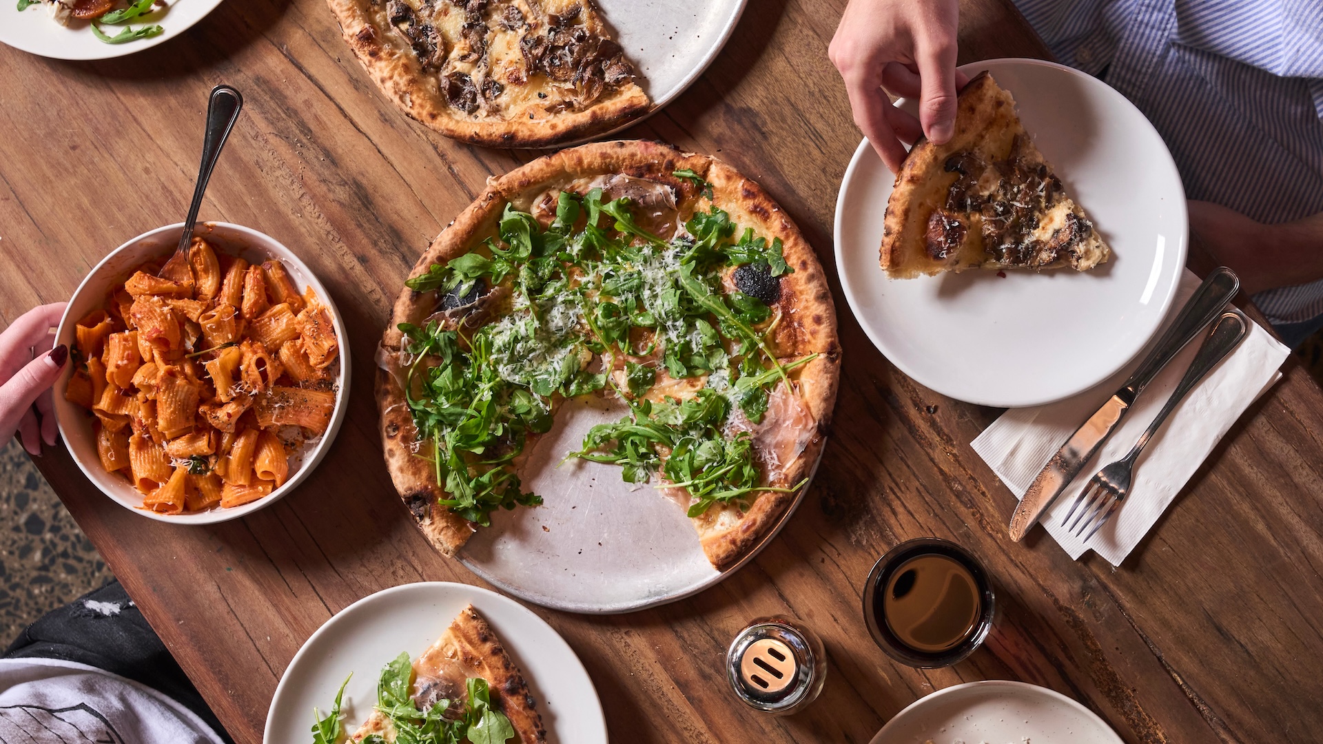 An overhead view of a rustic wooden table spread with Italian dishes, including a wood-fired pizza topped with arugula and shaved parmesan, a bowl of rigatoni in tomato sauce, and additional pizza slices on white plates. Two people's hands are visible reaching for food.