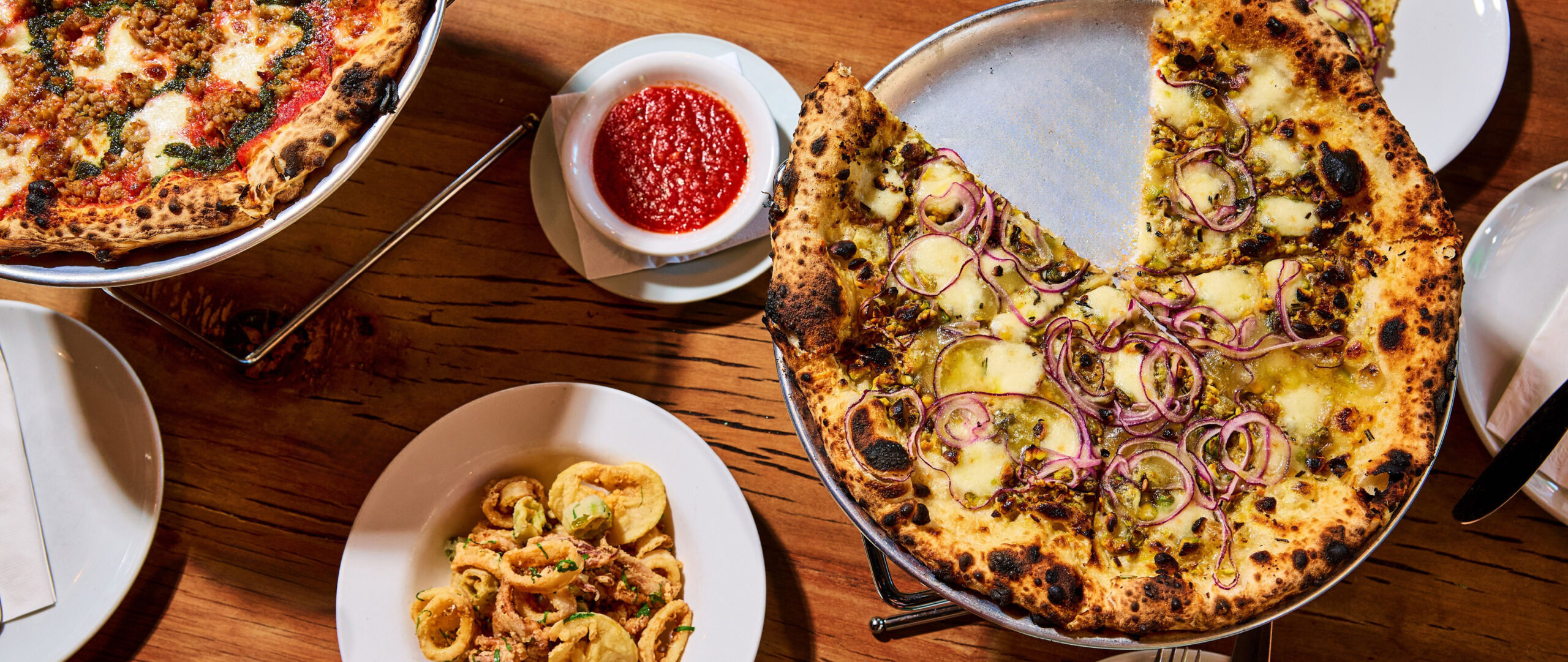 Overhead view of Pizzeria Stella wood-fired pizzas with pickled peppers and red onion and sausage with herbs, served alongside calamari fritte and marinara dipping sauce