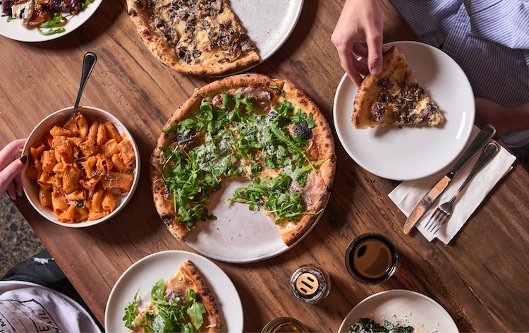 An overhead view of a rustic wooden table spread with Italian dishes, including a wood-fired pizza topped with arugula and shaved parmesan, a bowl of rigatoni in tomato sauce, and additional pizza slices on white plates. Two people's hands are visible reaching for food.