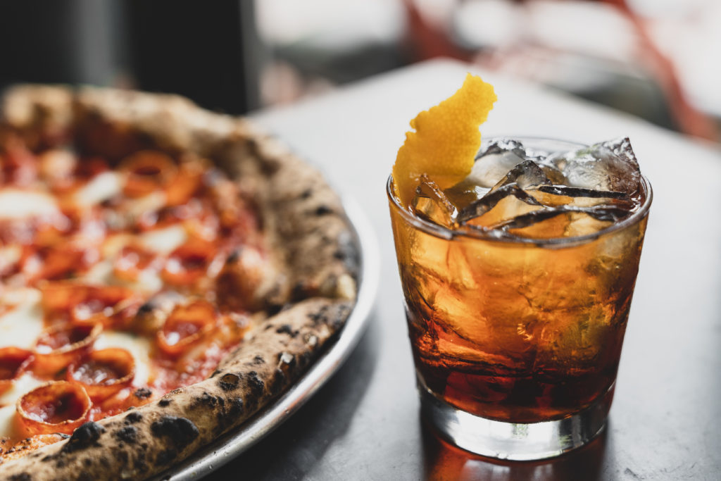 A pepperoni pizza and a drink with ice and an orange peel.